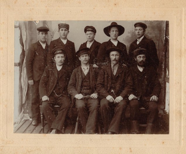 Picture of nine men, five standing, with four seated in front.
